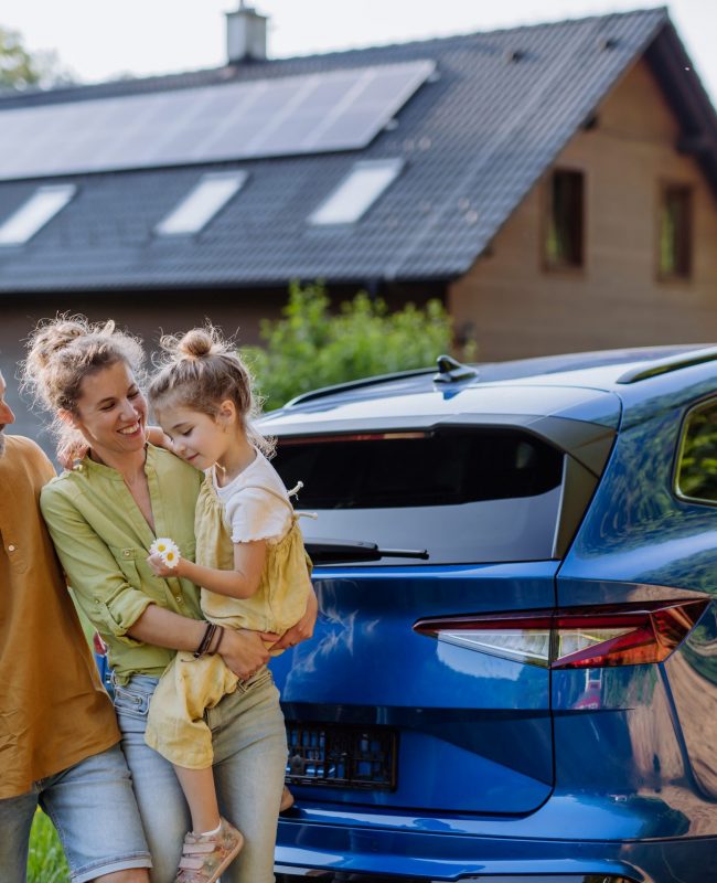 Family with little girl standing in front of their house with solar panels on the roof, having electric car. Family with little daughter standing in front of their house with solar panels on the roof, having electric car.